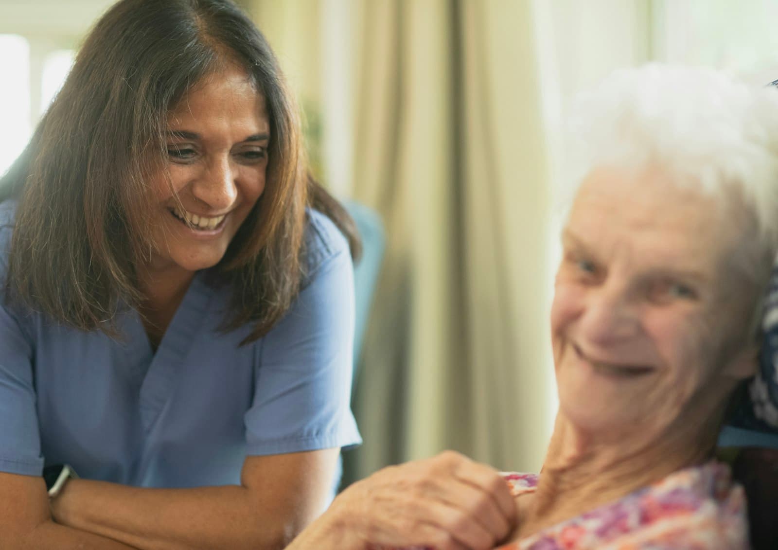 A caregiver speaking warmly with an older woman near a sunny window.
