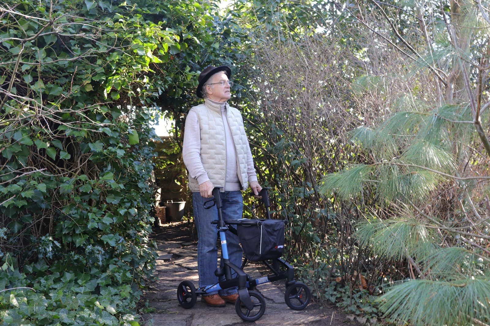 An older man standing outdoors with a rollator walker on a garden path.