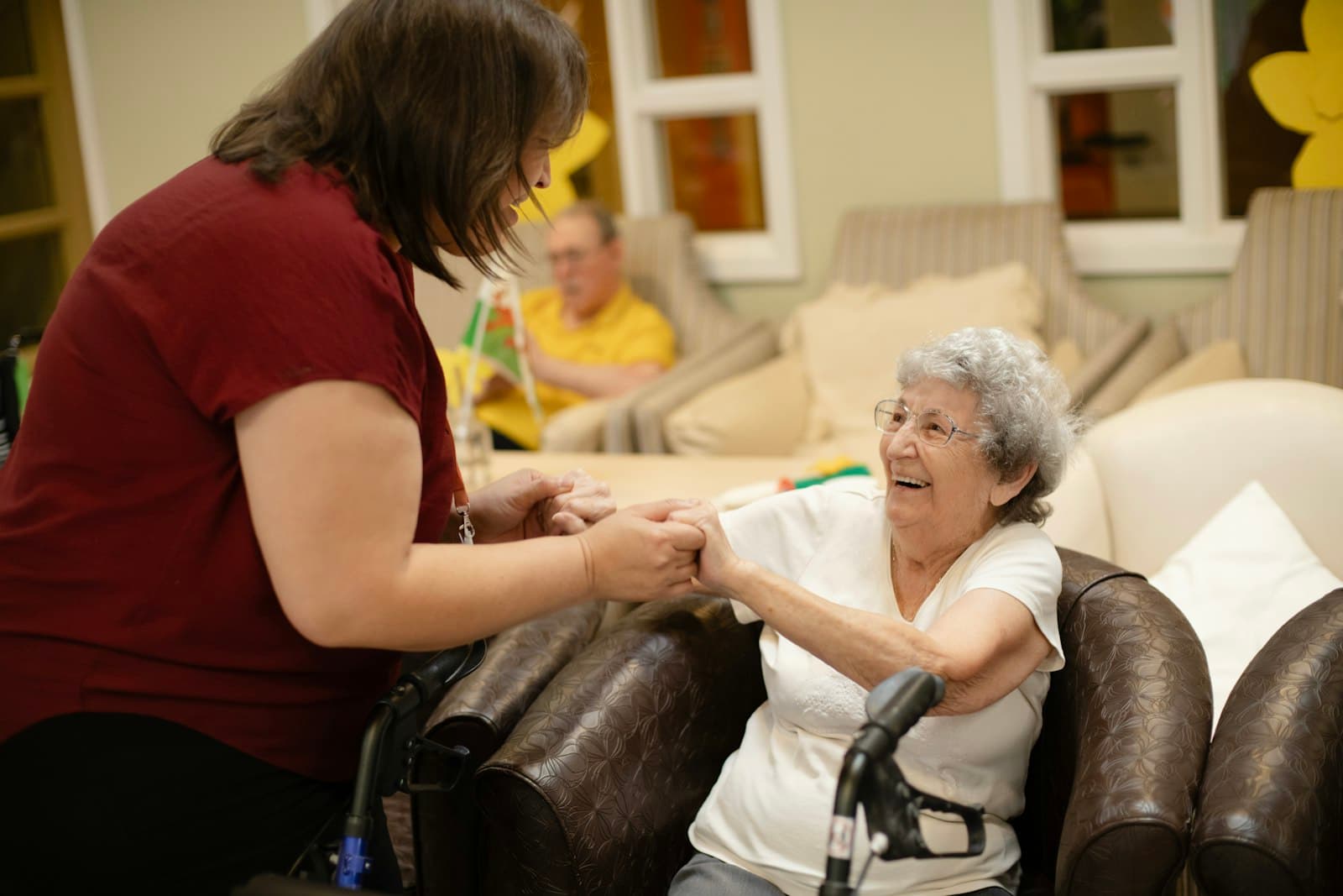 A caregiver helping an older woman rise from a chair while a walker sits nearby.